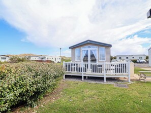 A mobile home with a fenced deck and a small porch, surrounded by a grassy area and a hedge. - The Retreat (Holywell)