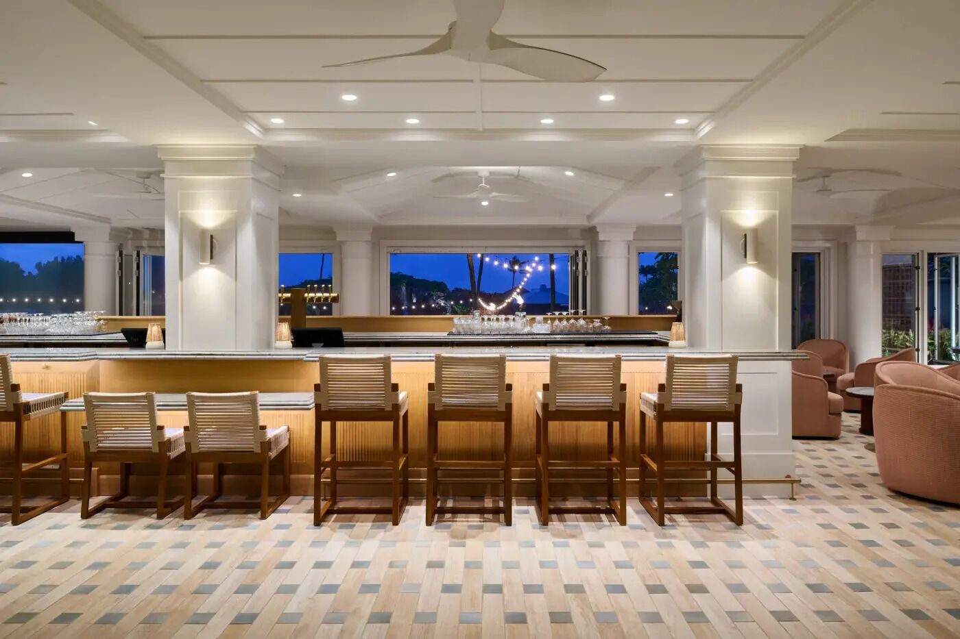 A bar area with a long counter, multiple bar stools, and a view of the city at dusk.
