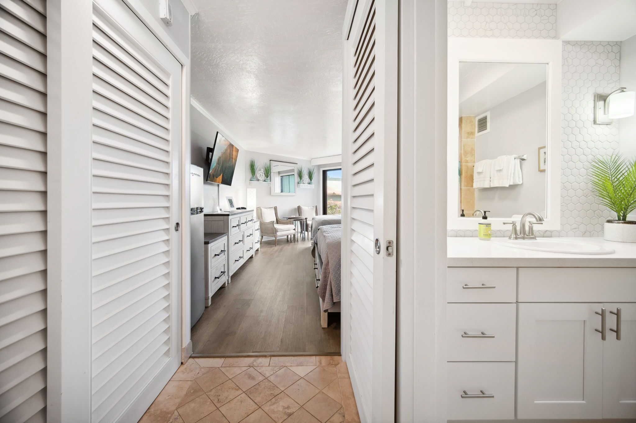 A modern, well-lit hallway leading to a bedroom with a large window, a bathroom with a sink and towel rack, and a kitchen area with cabinets and a countertop.