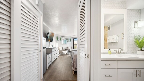 Un pasillo moderno y bien iluminado que conduce a un dormitorio con un ventanal grande, un baño con lavamanos y perchero, y una zona de cocina con armarios y encimera.