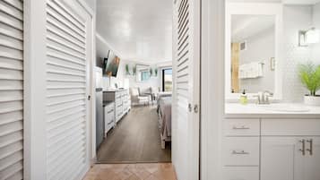 A modern, well-lit hallway leading to a bedroom with a large window, a bathroom with a sink and towel rack, and a kitchen area with cabinets and a countertop.