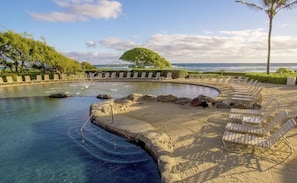 A pool with clear water, surrounded by lounge chairs and a palm tree, with a view of the ocean.