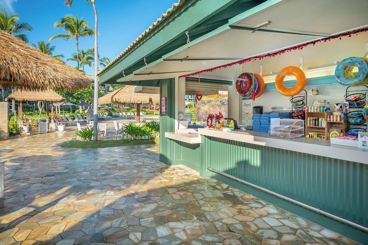 Un petit kiosque situé en bord de plage, doté d’une terrasse carrelée en pierre, d’une structure à toit de chaume et proposant une variété d’articles à la vente.