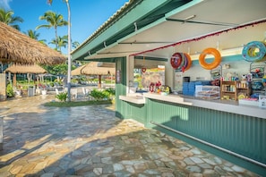 A beachside kiosk with a stone-tiled patio, a thatched-roof structure, and a variety of items for sale.