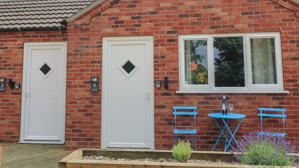 Une maison à deux étages, construite en briques, avec deux portes blanches, une fenêtre offrant une belle vue et un petit espace extérieur aménagé avec des chaises bleues.