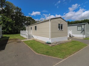 A single-story mobile home with a front porch and a small deck, surrounded by a grassy area and trees. - Forest Retreat - Fo26 (Saxmundham)