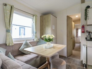 A compact living space with a sofa, a small table with a vase of flowers, a kitchenette, and a door leading to another room. - Forest Retreat - Fo26 (Saxmundham)