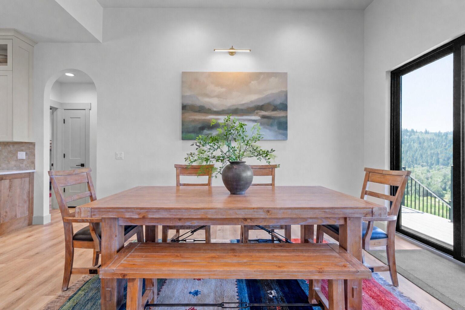 A dining area with a wooden table, chairs, a vase with greenery, and a painting on the wall.
