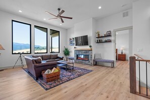 A spacious living room with a large window, a fireplace, and a ceiling fan. - Brand-New Mountain Retreat Sleeps 8 (Palisades)