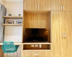 Ein Zimmer mit an der Wand montiertem Fernseher auf einer Holdecke, einem petrolfarbenen Sessel und einem Regal mit gerahmten Bildern.
