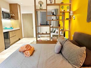 Ein Schlafzimmer mit Bett, Kissen, einem Holzregal und Blickrichtung auf eine Küche und ein Badezimmer.