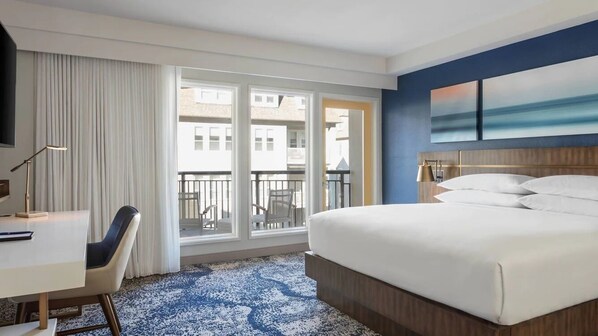 Ein Hotelzimmer mit Bett, Schreibtisch, Stuhl, Fernseher und einem Balkon mit Blick auf Gebäude.