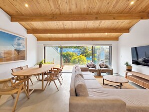 Ein modernes Interieur mit Holzbalkendecke, einem Essbereich mit Holztisch und -stühlen, einer Couch und einem an der Wand befestigten Fernseher.