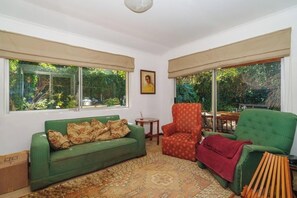 Een kamer met een groene bank, een rode fauteuil, en een groene fauteuil met een rode plaid. Er zijn twee ramen met beige gordijnen, en een schilderij aan de muur.