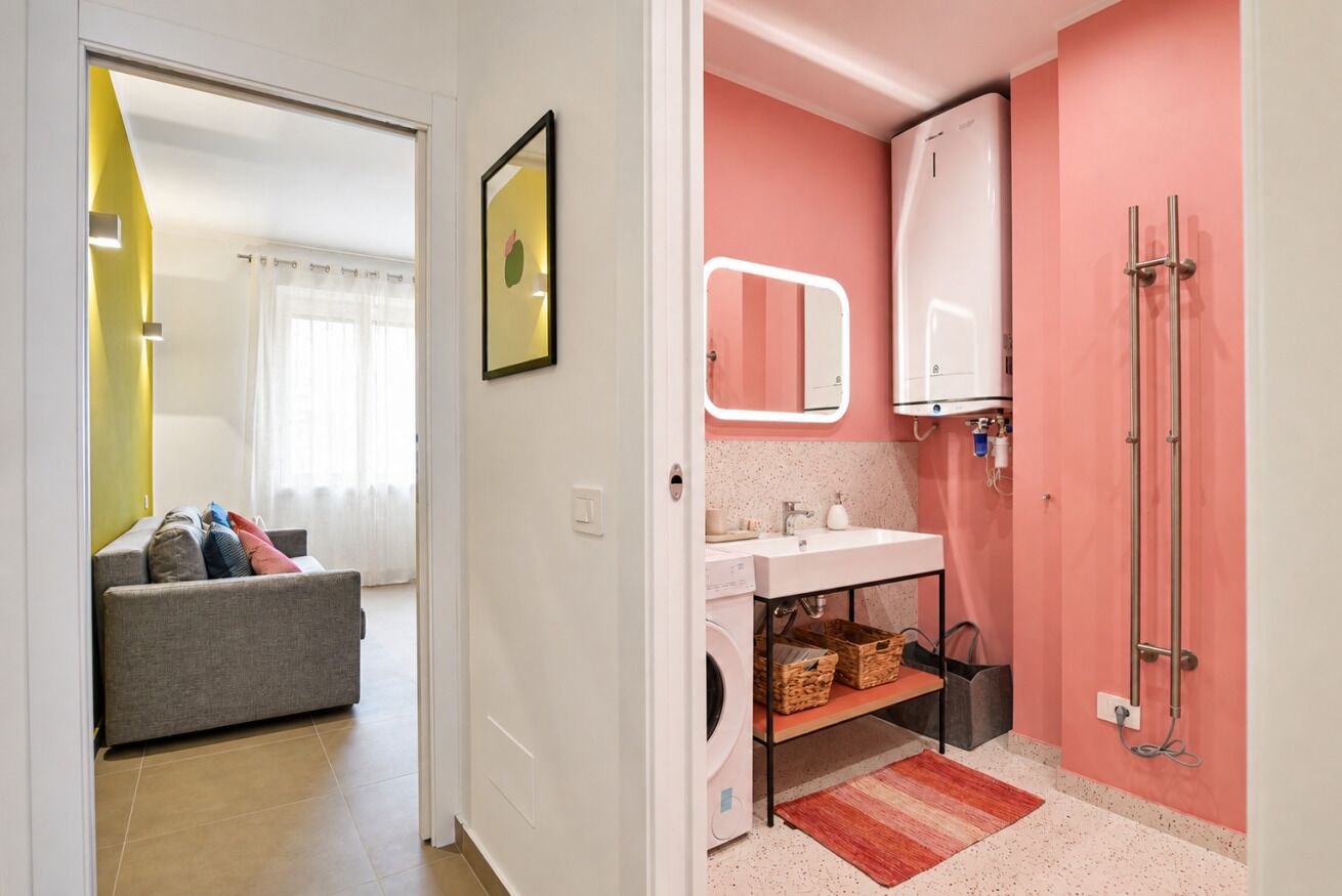 A modern bathroom with a pink wall, a white sink, a mirror, a shower, and a laundry area.