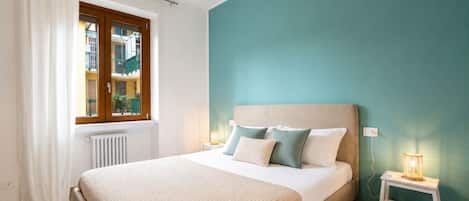 A bedroom with a bed, a window with curtains, a bedside lamp, and a view of a building outside.
