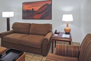 Ein Wohnzimmer mit einer Couch, einem gestreiften Sessel, einer Lampe, einem kleinen Tisch mit Telefon und Unterlagen sowie einem Gemälde einer untergehenden Sonne.