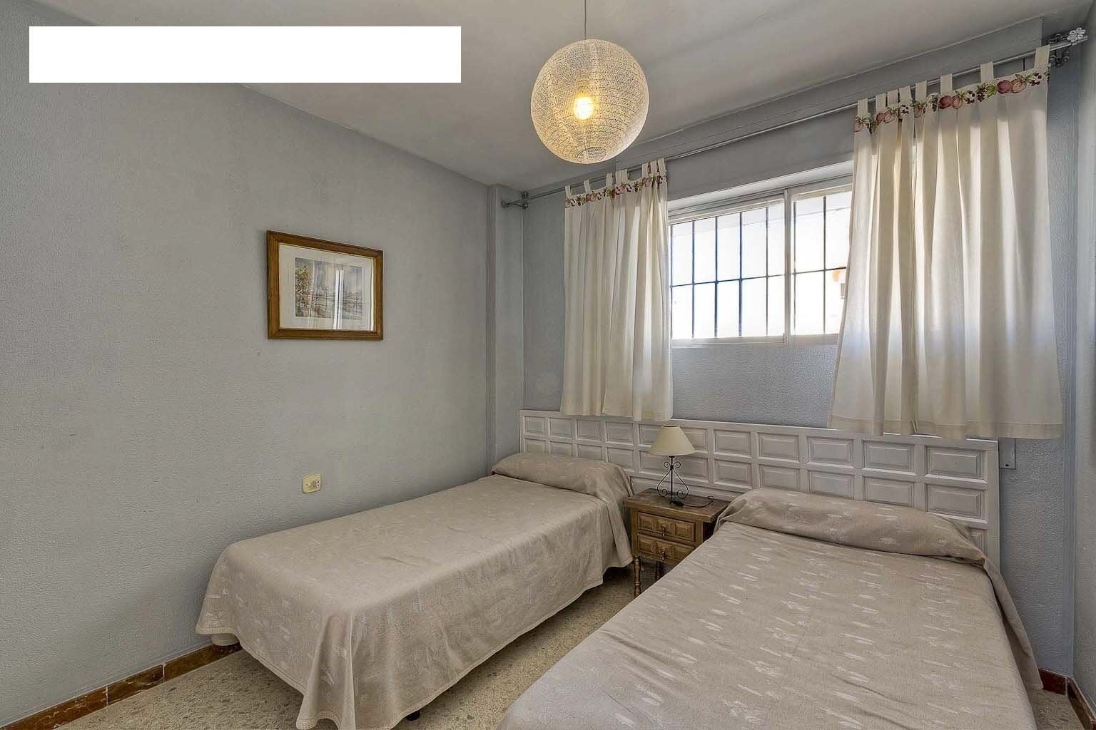 A room with two beds, a hanging light fixture, a window with curtains, and a framed picture on the wall.