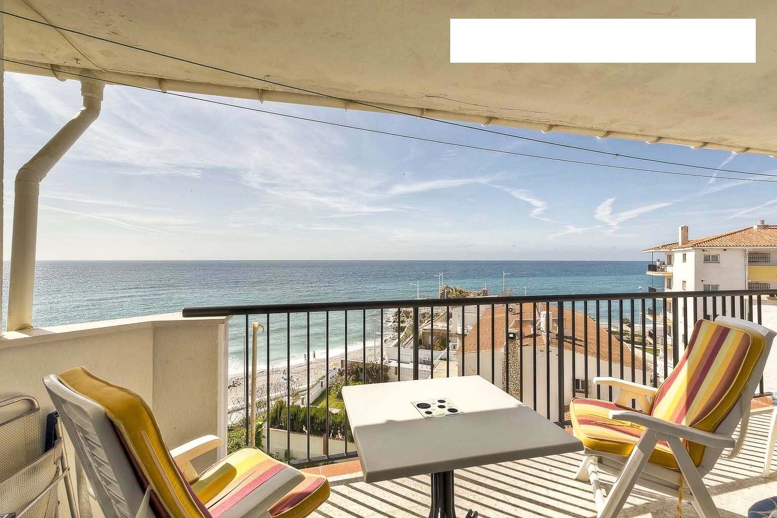 A balcony with a view of the sea, a table, and striped chairs.