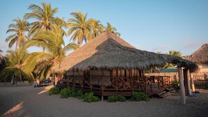 A thatched-roof hut with a wooden deck, surrounded by palm trees and a sandy beach.