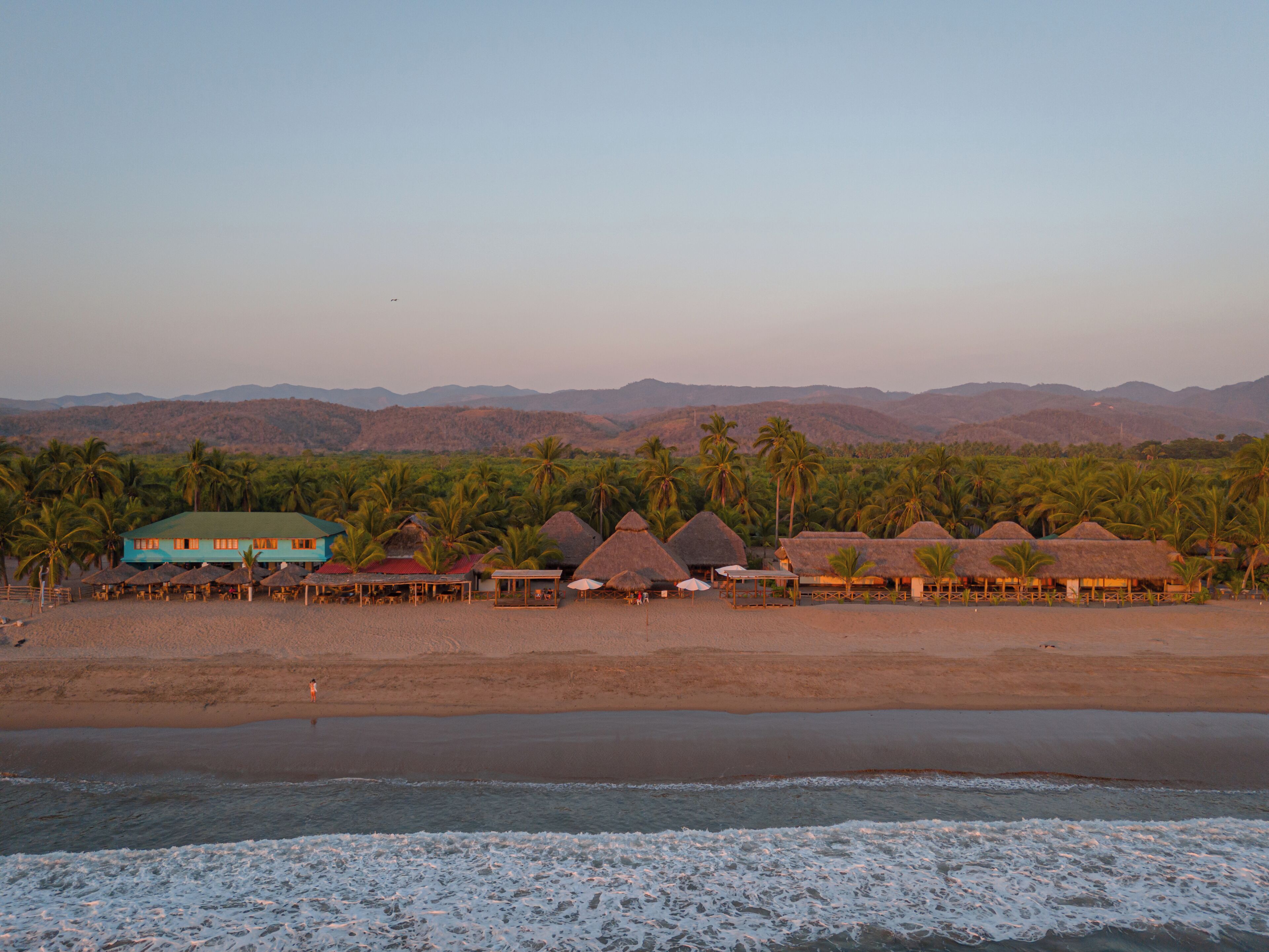 A beachfront property with thatched-roof huts, palm trees, and a mountain backdrop.