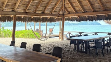 En hytte ved stranden med stråtækt tag, træbjælker og et spisebord med stole.