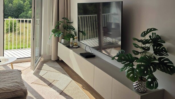 A modern living room with a flat-screen TV mounted on a white cabinet, a sofa, a coffee table, and a large window with curtains.