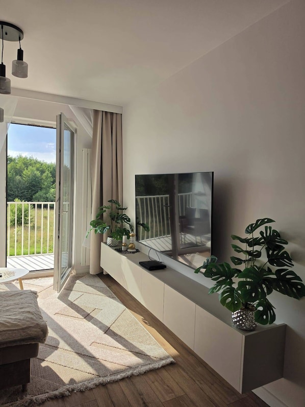 A modern living room with a flat-screen TV mounted on a white cabinet, a sofa, a coffee table, and a large window with curtains.