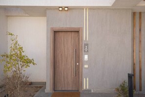 A modern building entrance with a wooden door, a number plaque, and a small plant to the left.