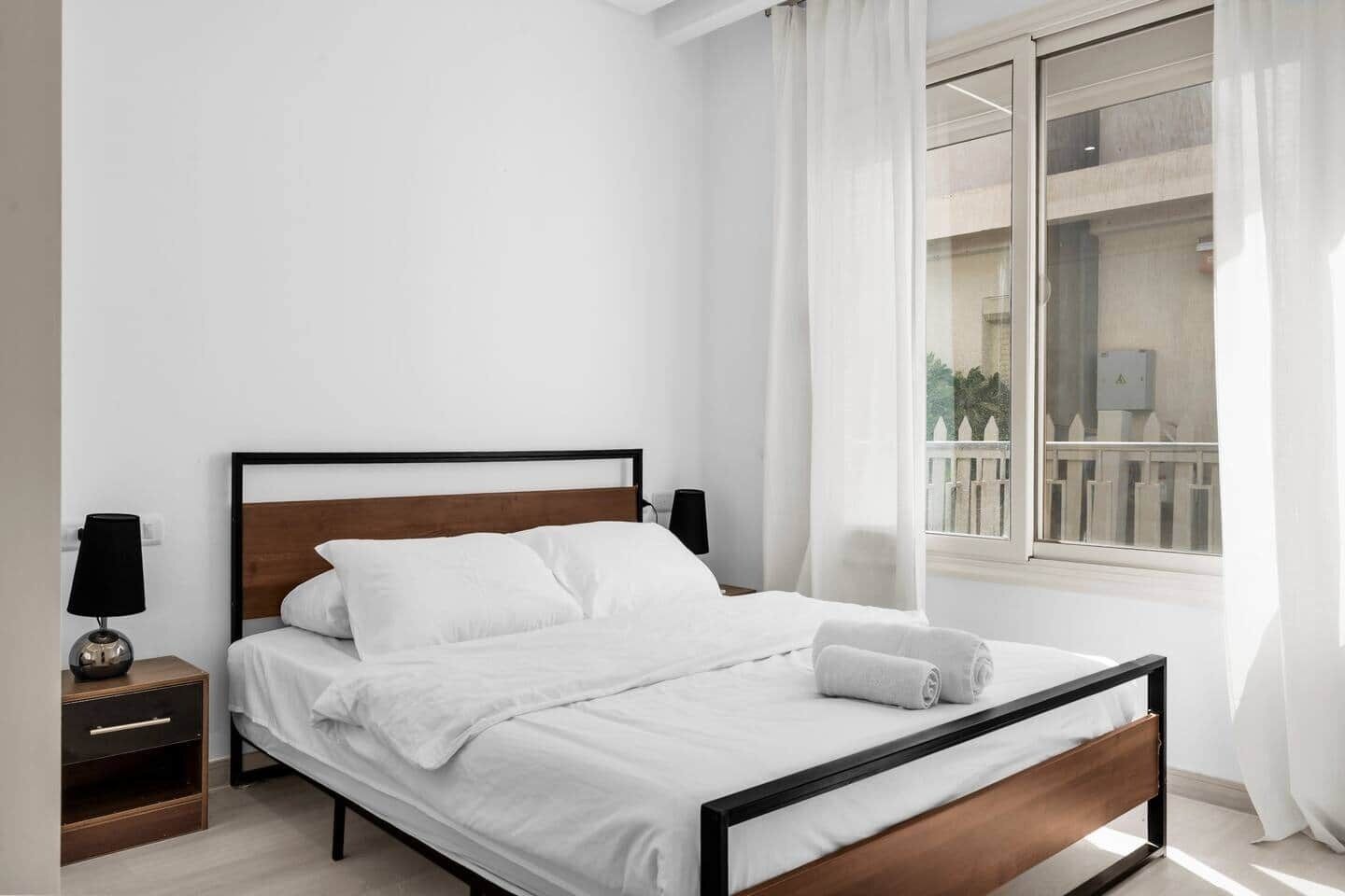 A neatly made bed with white linens and a wooden headboard in a room with white walls and a window with curtains.