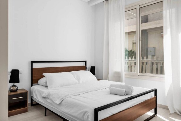 A neatly made bed with white linens and a wooden headboard in a room with white walls and a window with curtains.