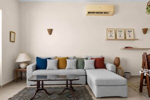 Ein Wohnzimmer mit grauem Ecksofa, einem Couchtisch, einer Lampe, einem Teppich, einer Klimaanlage, Wandleuchten, gerahmten Kunstwerken und einem Bücherregal.