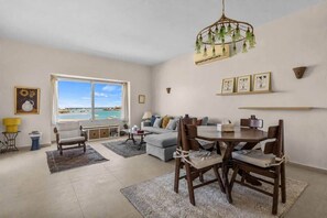 A spacious living room with a large window overlooking a beach, a dining area with a wooden table and chairs, and a cozy seating area with a sofa and armchairs. - The Sandy Feet. Part II - Nesty Hospitality (Hurghada)