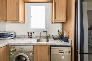 A compact kitchen with wooden cabinets, a built-in washing machine, a dishwasher, and a window above the sink. - The Sandy Feet. Part II - Nesty Hospitality (Hurghada)