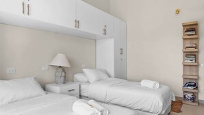 A hotel room with two beds, a bedside table with a lamp, a wardrobe, and a bookshelf. - The Sandy Feet. Part II - Nesty Hospitality (Hurghada)