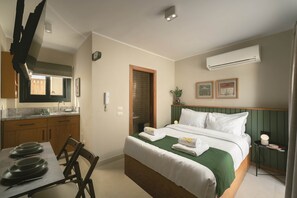 A hotel room with a bed, a small bedside table, a chair, a dining table with plates, a window, and an air conditioning unit. - Solé Verde Rooftop Studio I Nesty Hospitality (Al Maadi)