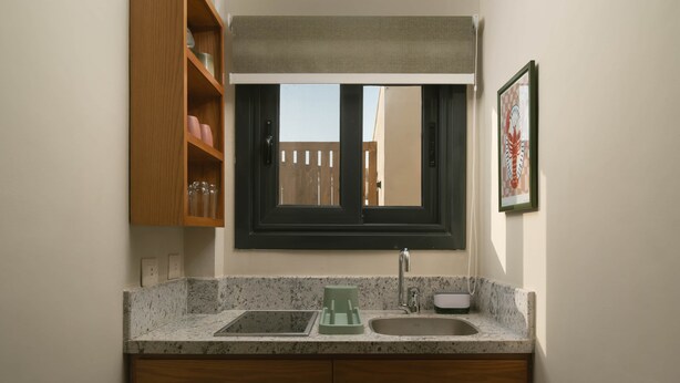 A kitchen with a double sink, wooden cabinets, a window with a view, and a framed picture on the wall. - Solé Verde Rooftop Studio I Nesty Hospitality (Al Maadi)