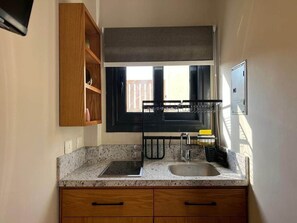 A compact kitchen with a granite countertop, a double sink, wooden cabinets, and a window with a black blind. - Solé Verde Rooftop Studio I Nesty Hospitality (Al Maadi)