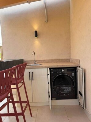 A compact kitchen with a built-in washing machine, a sink, and a bar area with stools. - Solé Verde Rooftop Studio I Nesty Hospitality (Al Maadi)
