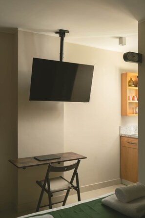 Ein Zimmer mit Fernseher an der Wand, einem Schreibtisch mit Laptop, einem Stuhl und einer kleinen Küchenzeile mit Schrank und Arbeitsplatte.