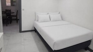 Habitación con una cama individual, una colcha blanca, una pequeña mesa con sillas y un ventanal.