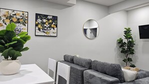 Ein modernes Wohnzimmer mit grauem Sofa, einem Esstisch, einer Kübelpflanze und zwei gerahmten floralen Gemälden an der Wand.