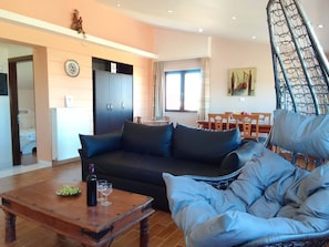 A living room with a sofa, a coffee table with a bottle and glasses, a dining area with chairs and a table, and a staircase with a metal railing. - Sofita Di Sofo Kos Vacation Apartment (Kos)
