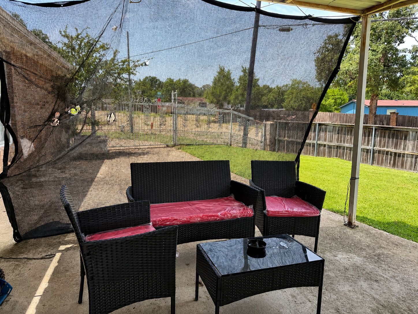 An outdoor patio with wicker furniture and a glass table, surrounded by a fenced yard and a clear sky.