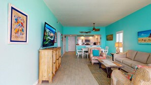 A living room with a television, a sofa, a dining area with chairs, and a kitchen in the background. - Beachside 2 4370 Coastal Living decor direct beach access pool (Miramar Beach)