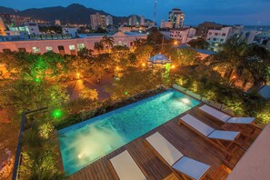 Ein Pool auf dem Dach mit Holzterrasse und Liegestühlen, umgeben von Bäumen und Stadtgebäuden.