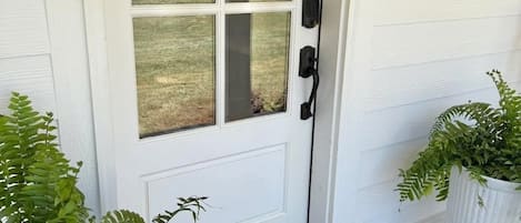 A white door with a glass panel, a black doorbell, a welcome mat, and two potted plants.