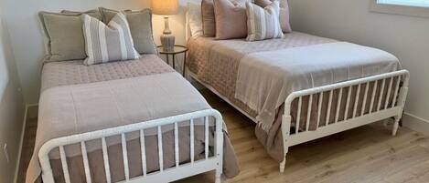 Two white metal bunk beds with a wooden floor, a lamp, and a window with blinds.
