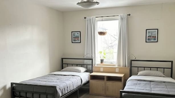 A room with two single beds, a window with curtains, a small table, and a wall-mounted shelf.