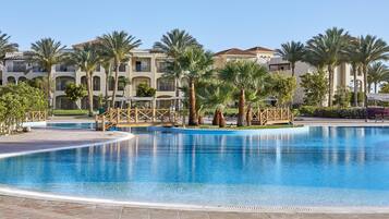A swimming pool with a wooden deck and palm trees, surrounded by a building with balconies.